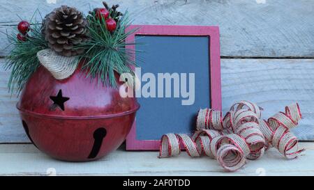 Campanello rosso rustico con stelle intagliate e cono di pino a forma di chiave, nastro verde e lavagna su uno sfondo di legno con spazio di scrittura Foto Stock