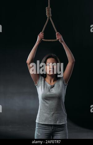 Premuto giovane donna andando a suicidarsi mentre si tiene la corda appesa cappio e guardando la fotocamera su sfondo nero Foto Stock