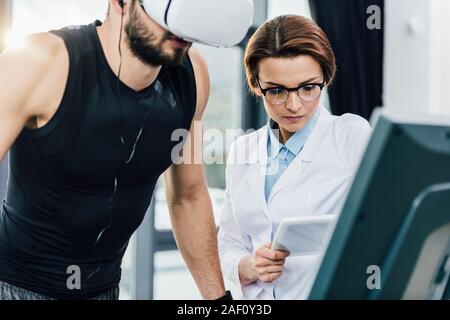 Sportivo in esecuzione sul tapis roulant vicino medico con tavoletta digitale durante la prova di resistenza nel centro sportivo Foto Stock