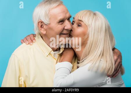 Senior donna baciando la guancia di allegro uomo con i capelli grigi isolato su blu Foto Stock