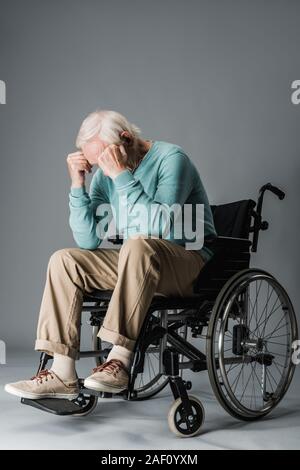 Ritirato uomo seduto nella sedia a rotelle e che ricopre la faccia su grigio Foto Stock