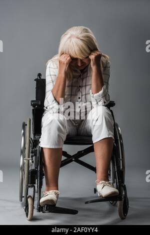 Sconvolto pensionati donna seduta in sedia a rotelle e toccando la testa su grigio Foto Stock