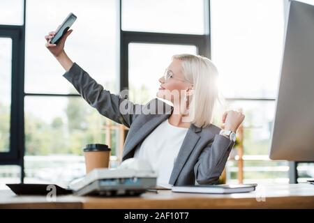 Messa a fuoco selettiva di allegro imprenditrice in bicchieri tenendo selfie in ufficio moderno Foto Stock