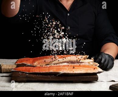 Chef in una camicia nera e nero Guanti in lattice prepara il filetto di salmone su un tagliere di legno, processo di aspersione con spezie e sale, chiave di basso Foto Stock