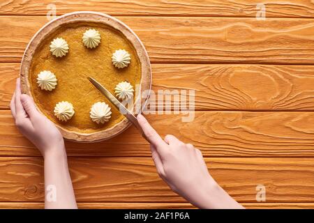 Vista ritagliata della donna il taglio di una deliziosa torta di zucca con panna montata su orange tavolo in legno Foto Stock