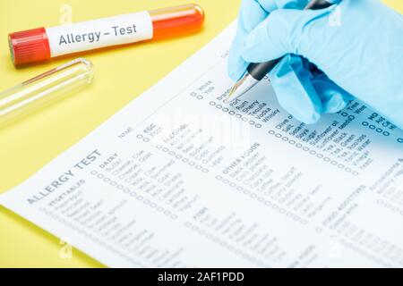 Vista ritagliata della donna nel guanto in lattice il riempimento in allergia modulo prova nei pressi di provette con allergia lettering di test isolato su giallo Foto Stock