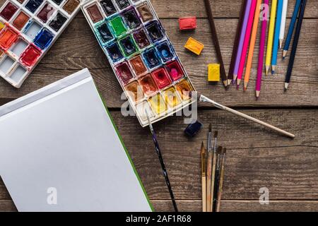 Vista superiore della vernice colorata di palette pennelli, matite di colore bianco e sketch pad su una superficie di legno Foto Stock