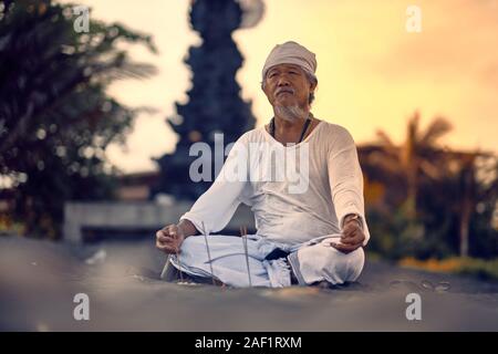 Bali, Indonesia - 10 DIC 2019: vecchio locale uomo balinese in meditazione vicino al mare. Concetto di pregare. Adulto pratica yoga sul Keramas spiaggia di sabbia nera Foto Stock