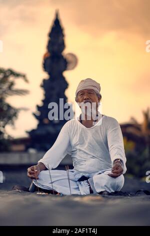 Bali, Indonesia - 10 DIC 2019: vecchio locale uomo balinese in meditazione vicino al mare. Concetto di pregare. Adulto pratica yoga sul Keramas spiaggia di sabbia nera Foto Stock