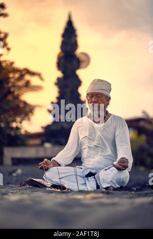 Bali, Indonesia - 10 DIC 2019: vecchio locale uomo balinese in meditazione vicino al mare. Concetto di pregare. Adulto pratica yoga sul Keramas spiaggia di sabbia nera Foto Stock