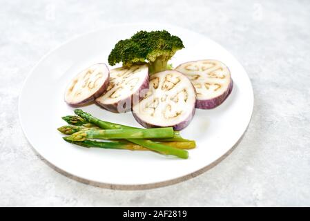 Verdure al vapore in una piastra bianca su un tavolo blu. Melanzana, Broccoli asparagi. Foto Stock