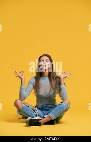 Vista frontale di una ragazza che mostra la pace utilizzando Lotus pongono. Elegante bella bruna donna seduta sul pavimento usando le dita per mostrare equilibrio, rendendo le labbra di anatra su sfondo giallo. Concetto di divertimento e di meditazione. Foto Stock