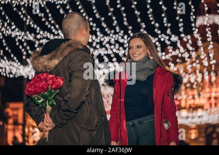 Il giorno di San Valentino o anniversario concetto - uomo nascondere un bouquet di rose dal suo sorpreso ragazza di notte in una strada di città Foto Stock