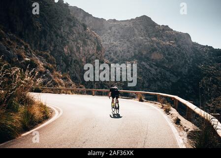 Da solo uomo sulla strada di bicicletta in salita in grandi montagne. Arancione tramonto luce in background Foto Stock
