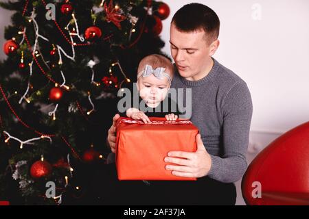 Bambina disimballaggio dono con il papà vicino albero di Natale.Felice ragazza spacchetta vacanze confezione regalo. Felice famiglia deliziosa Foto Stock