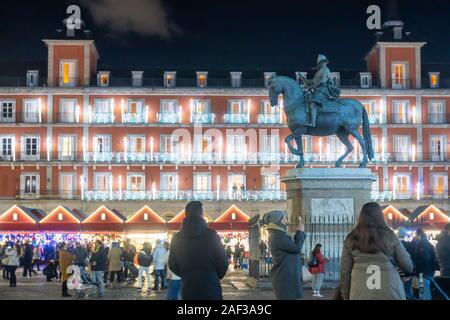 Una statua di re Filippo III di Spagna sorge in Plaza Mayor a Madrid durante la notte. Un mercato di Natale lo circondano la piazza è occupato con la gente. Foto Stock