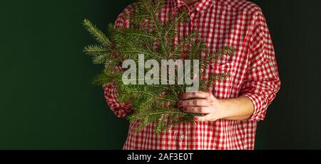 Una donna in una maglietta rossa è in piedi contro un muro verde, tenendo un albero di Natale ramo nelle sue mani e che copre il volto. Concetto di vacanza. Minimali Foto Stock