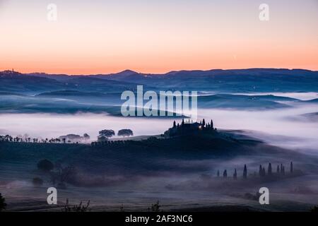 Tipico del territorio collinare della campagna toscana in Val d'Orcia, l'azienda agricola Podere Belvedere su di una piccola collina e la nebbia nelle valli, di sunrise Foto Stock