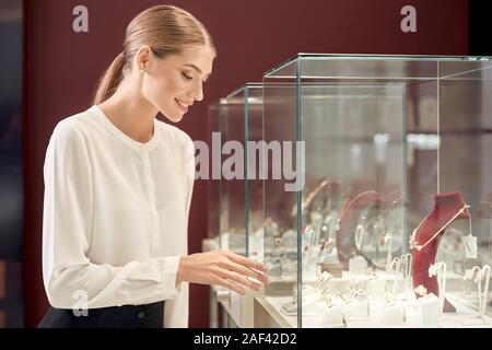 Bella donna con capelli biondi e legato il nodo in bianco seta camicetta sorridente e guardando in vetrina con gioielli di lusso in negozio. Assistente femmina controlla i l'aspetto appropriato di metalli preziosi. Foto Stock