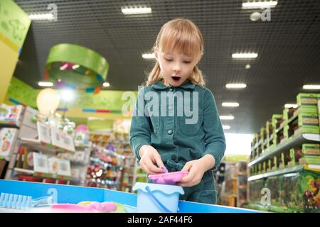 Ragazza Suprized in montagna con bocca aperta giocando con la plastilina nel grande negozio di giocattoli. Piccolo bambino femmina godendo, scelta e acquisto di nuovi giocattoli di fresco nel supermercato. Concetto di infanzia e di gioia. Foto Stock