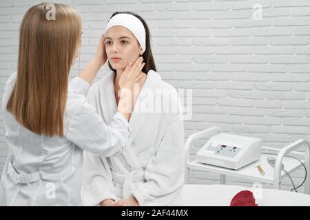 Vista frontale di grave donna seduta sul pullman mentre estetista femmina guardando la pelle del viso. Client di giovani preoccuparsi di bellezza, facendo rejuvenative e procedure mediche. Concetto di trattamento. Foto Stock