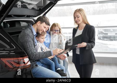 Vista laterale di femmine della concessionaria auto firma contratto con connettore maschio di tipo client in auto salon. Il cliente seduto in auto con la famiglia in show room. L'uomo con sua moglie e i bambini sorridenti scelta e acquisto di nuove automobili. Foto Stock