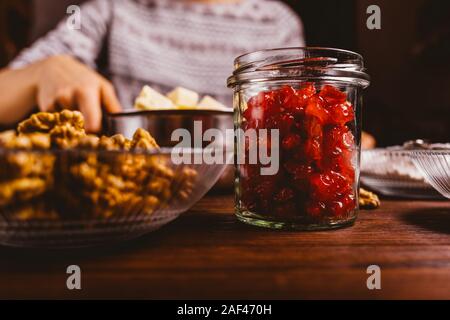 Immagine dello stile di vita processo di rendere dolci fatti in casa con ciliegie e dadi, close-up di frutti di bosco essiccati in vaso e noci in ciotola di legno sul tavolo da cucina Foto Stock