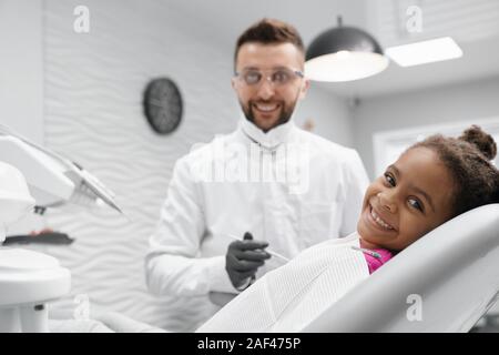 Vista dal lato di allegro bambino africano giacente sulla poltrona del dentista, guardando la fotocamera e sorridere mentre medico denti di indurimento. Dentista professionale mantenendo l'attrezzo e che pongono in background. Concetto di cura. Foto Stock
