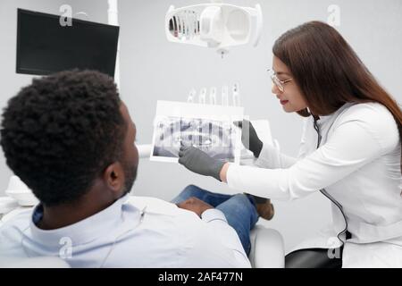 Vista dal lato della femmina attraente dentista mantenendo i raggi x immagine di denti e mostra paziente di sesso maschile in studio dentistico. Sorridente medico denti di indurimento in clinica. Concetto di stomatologia e medicina. Foto Stock