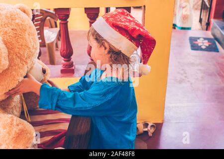 Allegro positivo bambina nel cappello di Natale e vestito blu è giocare con grande orso di peluche. Natale concetto di viaggio su un viaggio in luoghi tropicali. Foto Stock