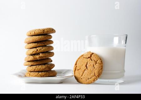 Un bicchiere di latte con un cookie di zenzero appoggiata contro di essa accanto a una pila di biscotti allo zenzero su una piastra bianca con uno sfondo bianco; spazio di copia Foto Stock
