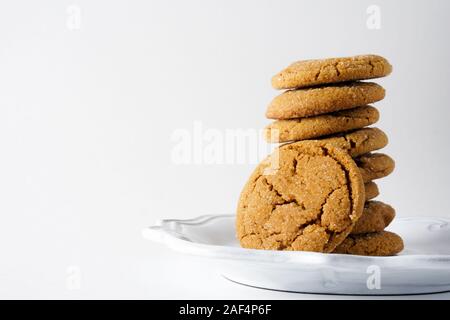 Un cookie di zenzero si appoggia contro una pila di biscotti allo zenzero su una piastra bianca e uno sfondo bianco con copia spazio sulla sinistra Foto Stock