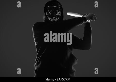 Ritratto di un anonimo uomo delinquente con la mazza da baseball in una felpa con cappuccio nero nasconde il suo volto dietro una maschera al neon. Studio shot. Foto Stock