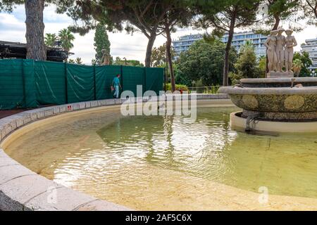 Una donna orologi come il suo piccolo cane gioca nell'acqua in corrispondenza delle tre Grazie fontana di Albert Park sulla Riviera Francese. Foto Stock
