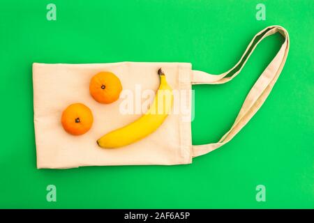 Tela di cotone borsa bianca e banana con i tangerini sul luminoso sfondo verde. Eco Friendly borsa riutilizzabile per lo shopping. Eco e rifiuti zero concetto. Foto Stock