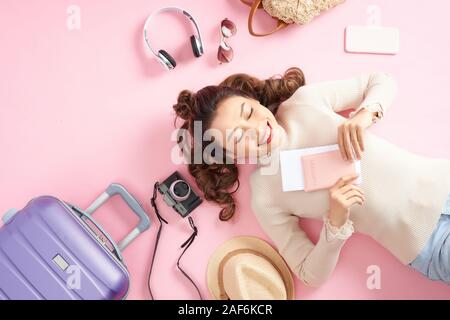 Donna asiatica tenendo il suo passaporto e giacente circondano il suo bagaglio di viaggio sul pavimento di colore rosa. Vista superiore Foto Stock