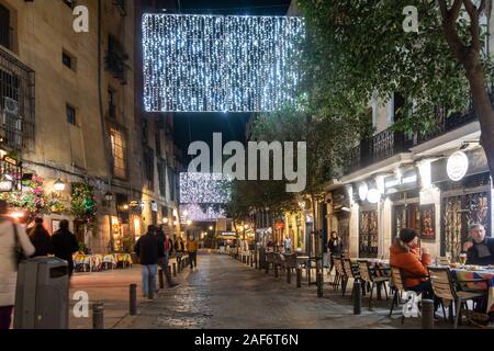 La gente seduta ai tavoli esterni a Cava de San Miguel a Madrid, Spagna. È Dicembre vicino a Natale e decorazioni che adornano le strade. Foto Stock