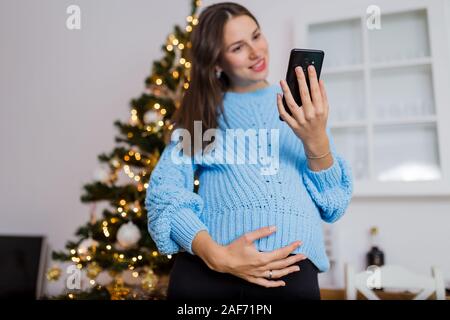 Ritratto di Allegro donna incinta utilizza lo smartphone accanto a albero di natale. Foto Stock