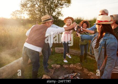 Gruppo di amici maturi rendendo Toast come essi siedono intorno al fuoco e cantare canzoni al campeggio all'aperto Foto Stock