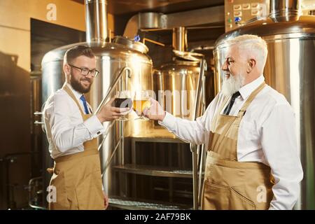 Due fabbricanti di esaminare e tenere la birra di bicchieri per la degustazione. Alcuni esperti in birreria vicino ad apparecchiature, canna. Il birraio e lavoratori in camice bianco, prua Foto Stock