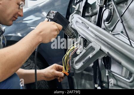 Vista laterale della macchina elettrica azienda aggiustatore e utilizzando il saldatoio per frullare i fili per la riparazione di segnalazione in porta smontata dell'auto. Serv Foto Stock