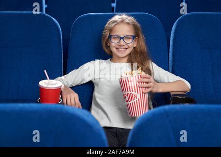 Vista frontale di felice ragazza in bicchieri guardando la fotocamera e ridere mentre si guarda la divertente commedia in cinema. Piuttosto bambino mantenendo il pop-corn e bere wa Foto Stock