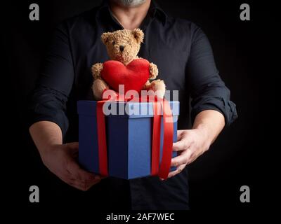 Un uomo adulto in una camicia nera contiene un quadrato blu box legate con un nastro rosso e si siede sulla parte superiore di un marrone orsacchiotto di peluche con un cuore, concetto di congratula Foto Stock