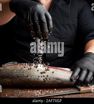 Chef in una camicia nera e nero Guanti in lattice prepara il filetto di salmone su un tagliere di legno, processo di aspersione con spezie e sale, chiave di basso Foto Stock