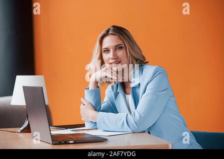 Se hai bisogno di consigli, mi può aiutare. Imprenditrice con ricci capelli biondi in ambienti chiusi in camera con colore arancio parete e tavolo in legno Foto Stock