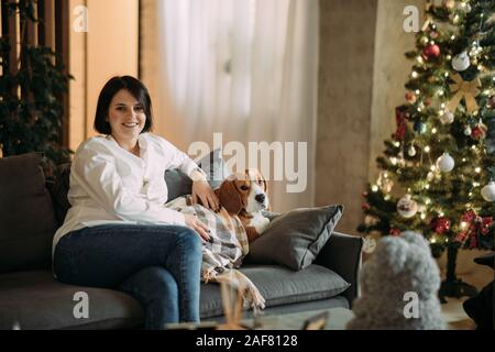 Una donna seduta su un divano accanto a un cane beagle e abbracci lui sullo sfondo di un albero di Natale. Foto Stock