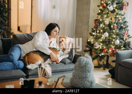 Una donna giace su un divano accanto a un cane beagle e abbracci lui sullo sfondo di un albero di Natale. Foto Stock