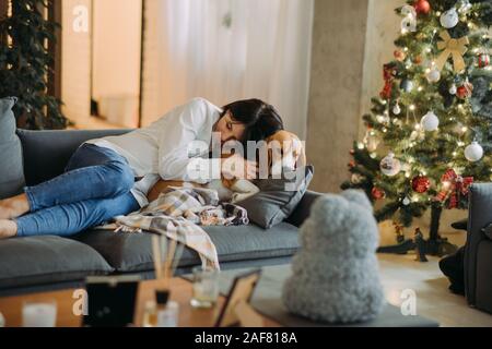 Una donna giace su un divano accanto a un cane beagle e abbracci lui sullo sfondo di un albero di Natale. Foto Stock