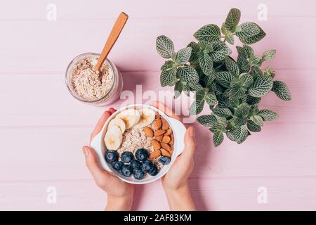 Femmina mani del recipiente di contenimento di deliziosi fiocchi d'avena con mirtilli, mandorle e banane accanto al vaso e pianta verde sulla tabella di rosa, vista dall'alto. Foto Stock