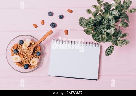 Il Granola sana colazione con noci e bacche accanto al notebook in bianco e verde houseplant sulla tabella di rosa, vista dall'alto Foto Stock